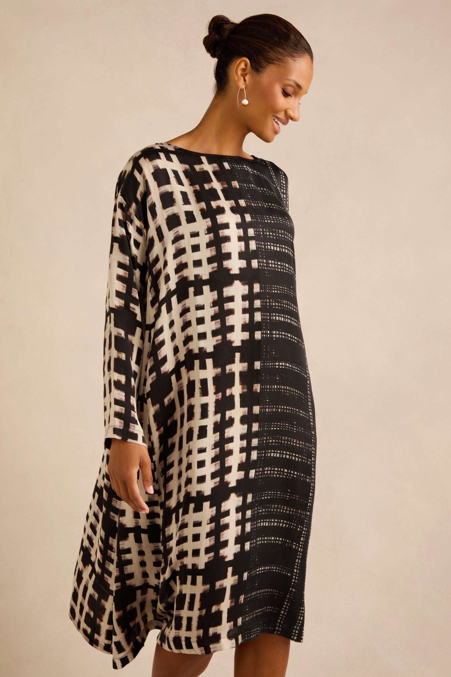 A woman wears the Check and Dash Easy Dress, featuring a black and white patterned long-sleeve, A-line silhouette, and side-swept design, standing against a plain beige background.