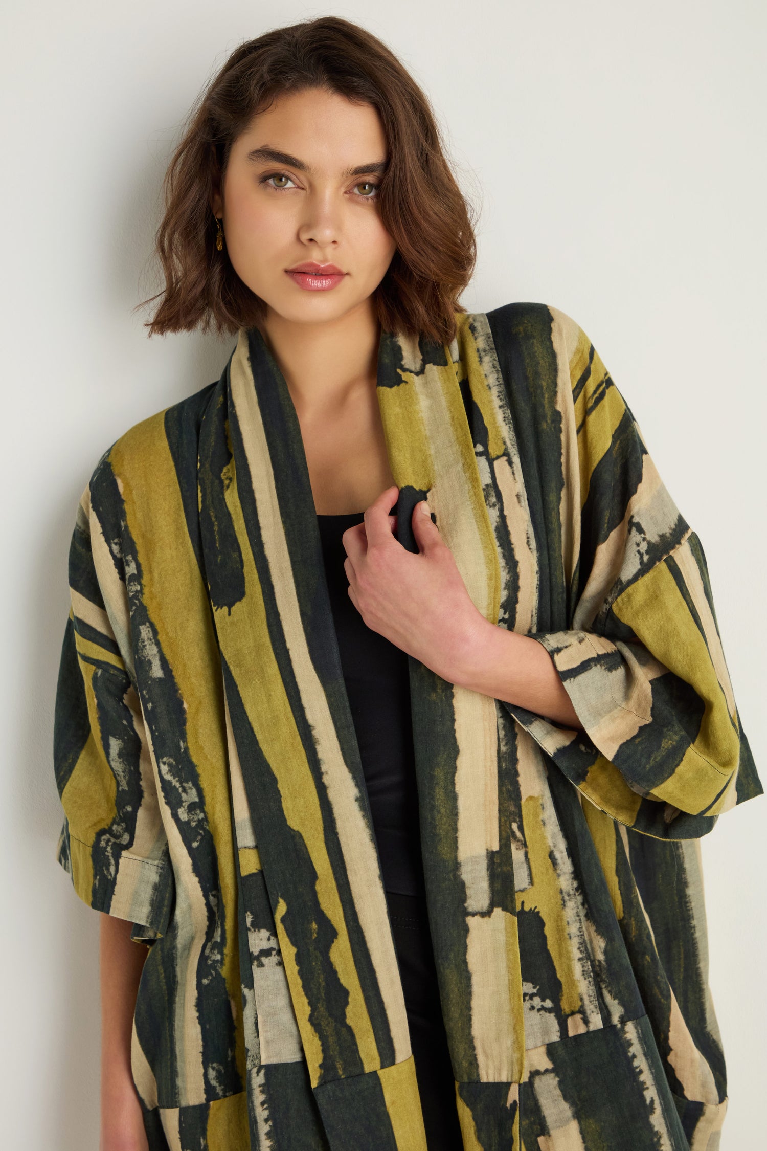 A woman with short brown hair wears a black top and the Oversized Cotton Kimono with a painterly yellow and dark green print, standing against a plain light background.