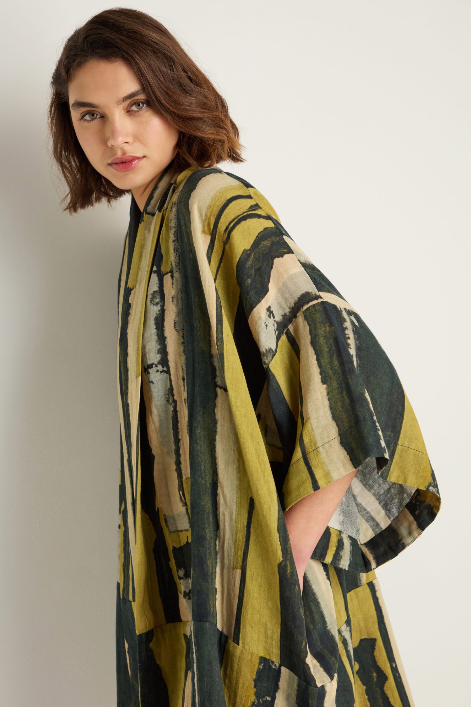 A woman with short brown hair models the Oversized Cotton Kimono, which features painterly green, yellow, cream, and black brushstroke prints, as she stands against a plain white wall.