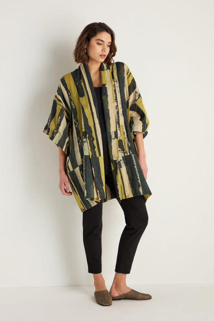 A woman stands against a white wall wearing the Oversized Cotton Kimono with a painterly print, paired with a black top, black pants, and brown slip-on shoes.