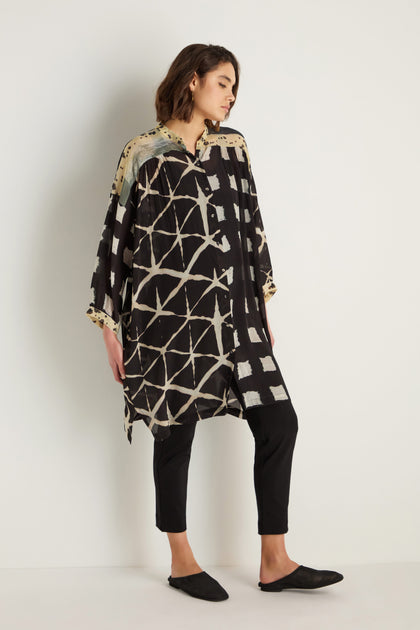 A person stands against a white wall wearing a relaxed-fit Shibori Print Long Shirt in black and beige, paired with black pants and black slip-on shoes.
