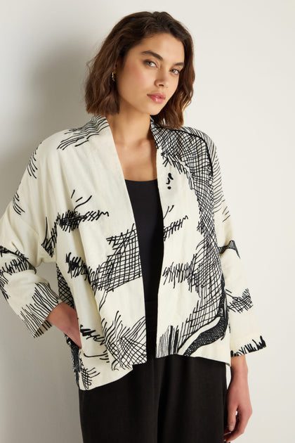 A woman stands against a plain light-colored wall wearing the Embroidered Sketch Jacket—an artisan-designed, white, open-front cotton jacket featuring black abstract line patterns—over a black top and pants.