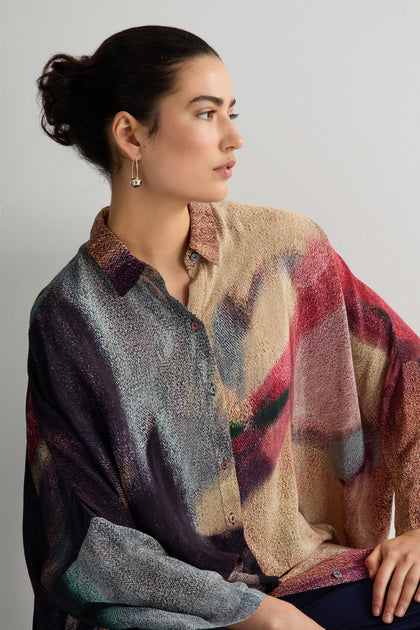 The individual with dark hair styled in a bun is wearing the Pixelated Shirt Dress with an abstract multicolored print, gazing to the side against a plain backdrop.