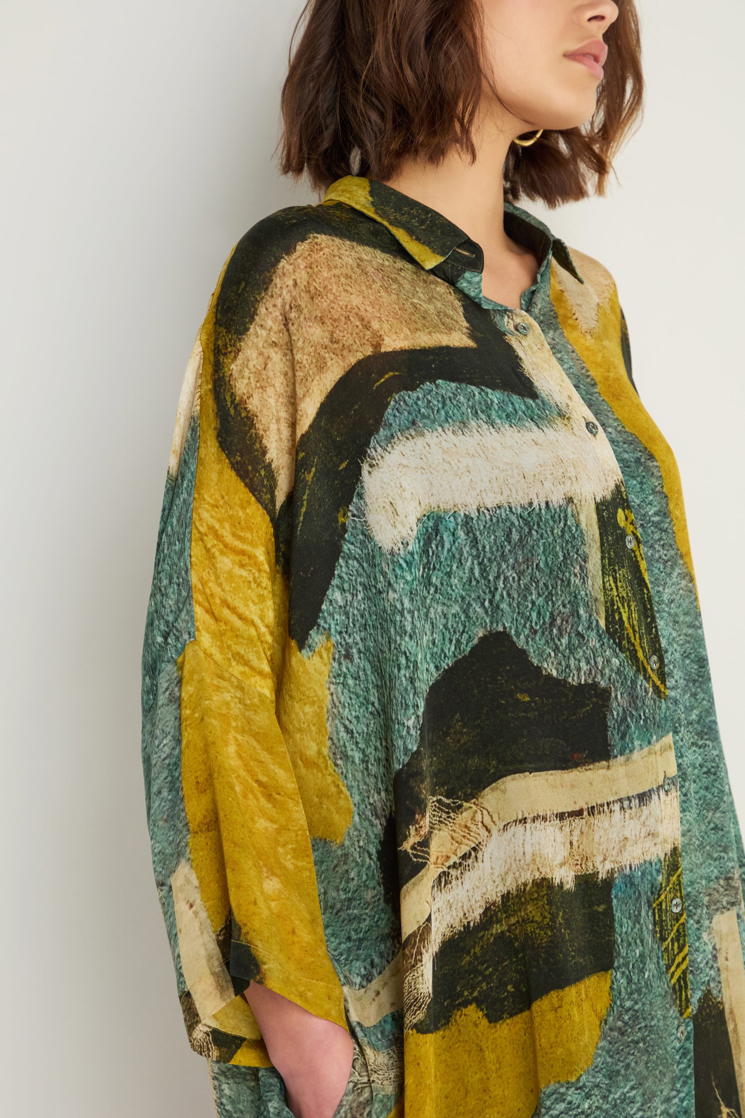 A woman models the Ochre Print Shirt Dress, featuring a loose fit and painterly green, yellow, white, and black prints. She stands against a plain background with her hands in her pockets.