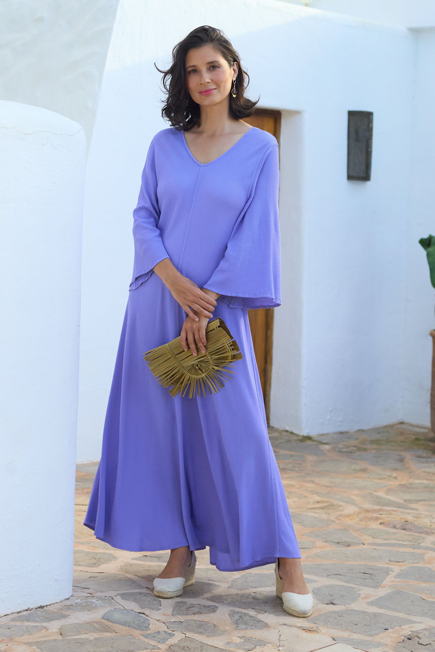 A woman in a Lilac Crepe Maxi Dress and cream shoes holds a gold clutch, standing confidently by a white wall.