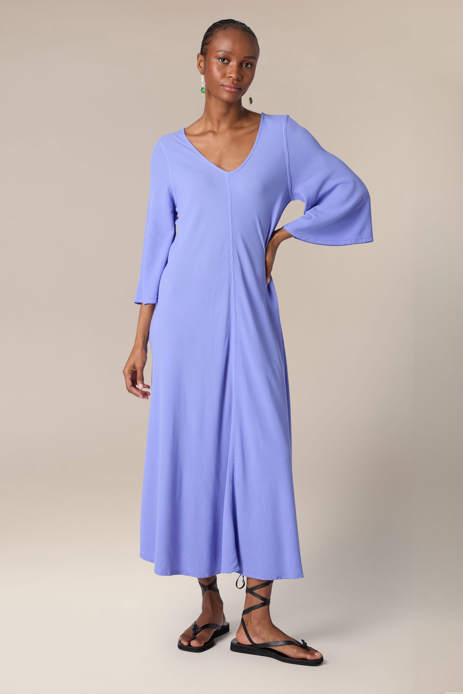 A woman wearing a stylish long blue Crepe Maxi Dress with three-quarter sleeves and black sandals stands posing against a beige background.