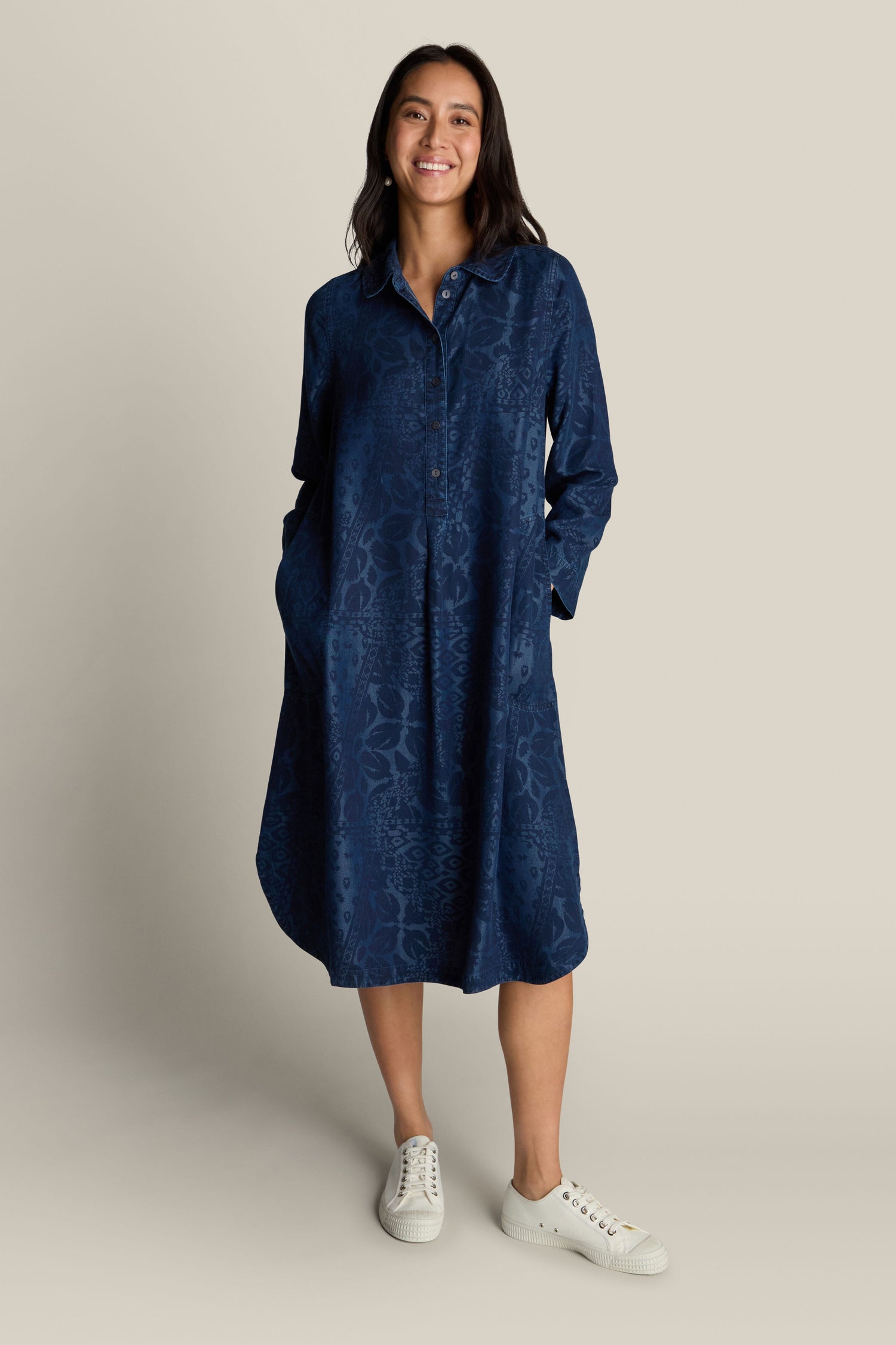 A person stands smiling, wearing the Abstract Patchwork Print Shirt Dress, a long-sleeve, button-up, knee-length blue dress with pockets, paired with white sneakers; the outfit showcases an abstract patchwork print for versatile styling options.