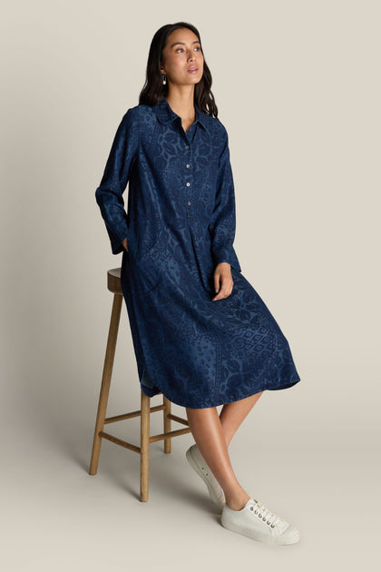 A woman in a long-sleeved, blue Abstract Patchwork Print Shirt Dress sits on a wooden stool, looking off-camera. She wears white sneakers and has her hands in the pockets of her versatile styling options dress.