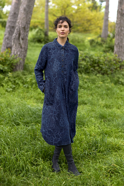 A person stands on grass in front of trees, wearing the Abstract Patchwork Print Shirt Dress styled with knee-high black boots.