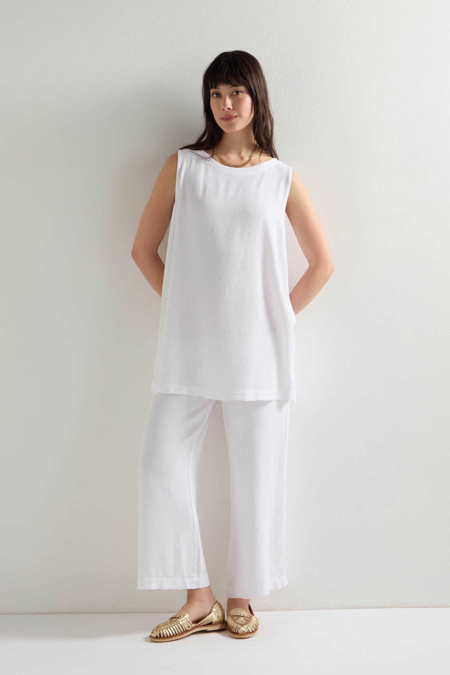 Dressed in a sleeveless white tunic, a person stands against a white wall wearing Crepe Wide Trousers with an elasticated waistband, combining comfort and elegance.