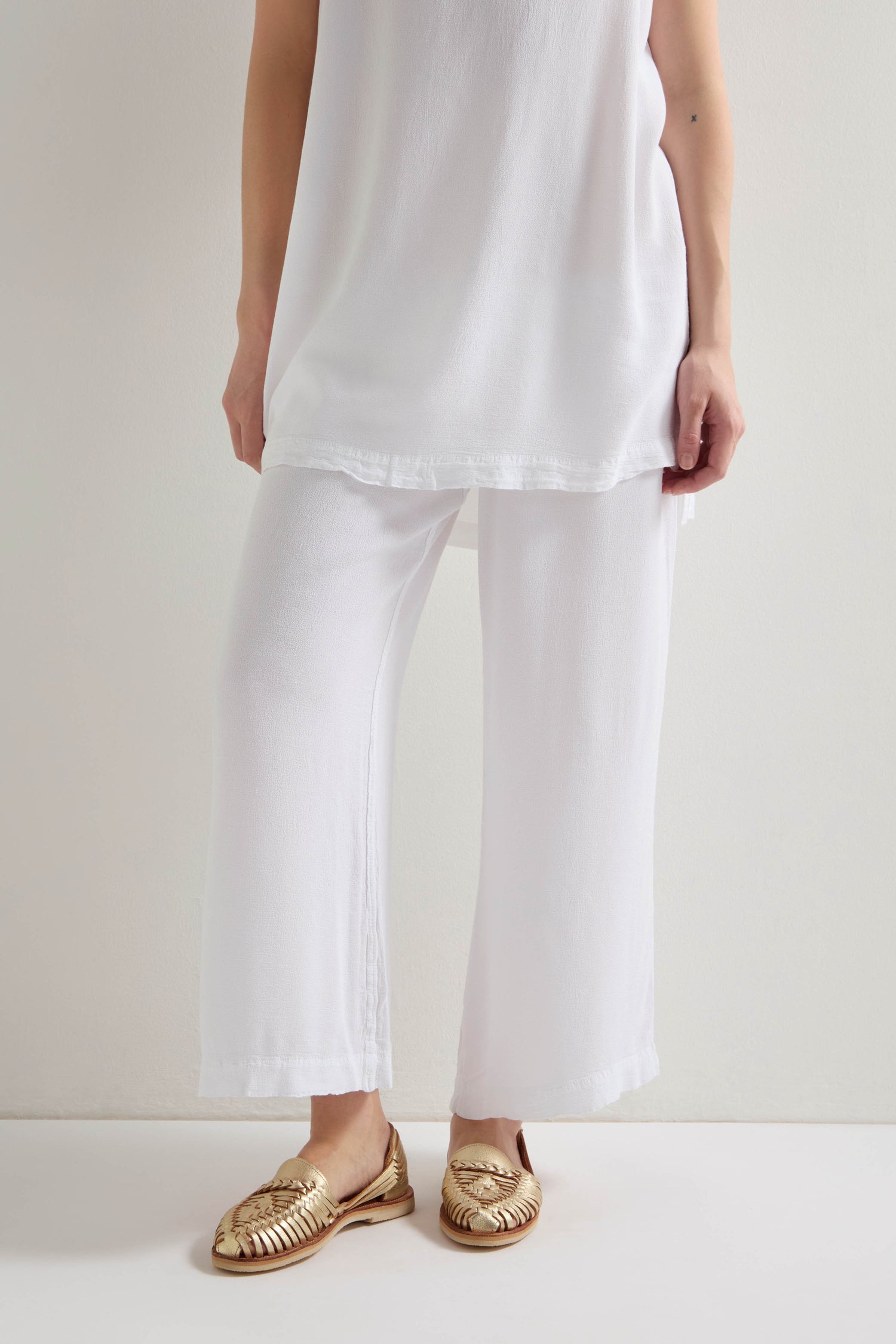 A person wears Crepe Wide Trouser in white, featuring a wide-leg silhouette and an elasticated waistband, paired with a matching top and gold woven sandals, standing against a light-colored wall.