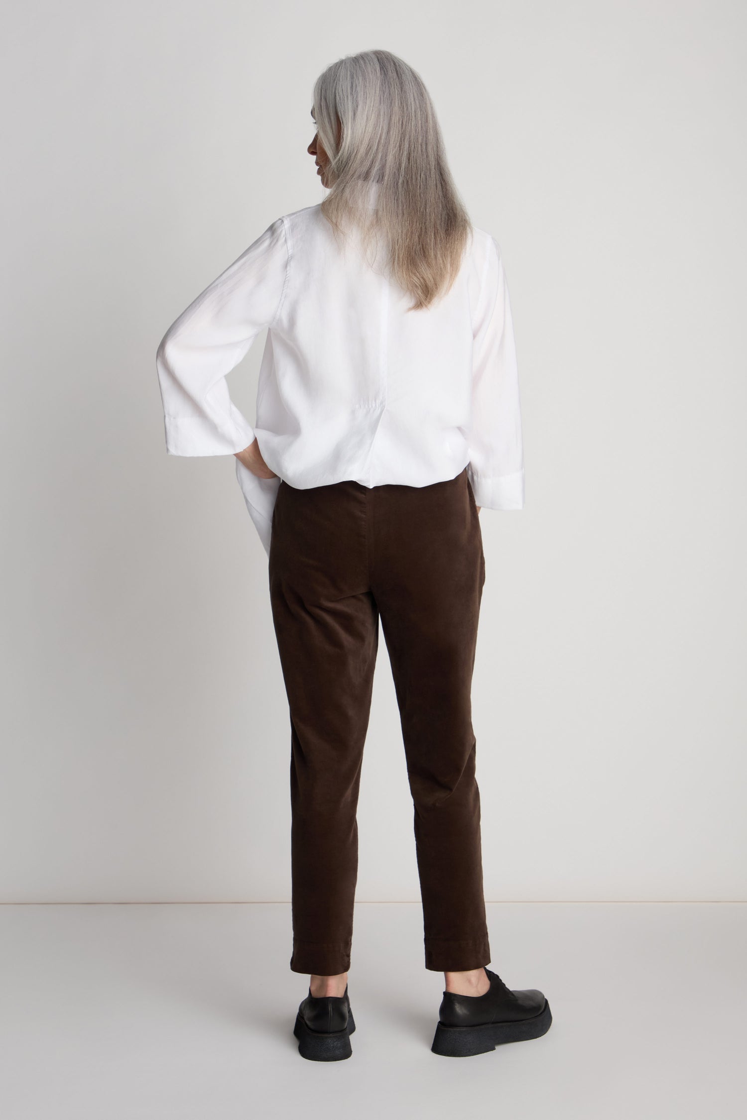 Rear view of a person with long gray hair wearing a white shirt, Cotton Moleskin Slim Trousers, and black shoes, standing against a plain white background.