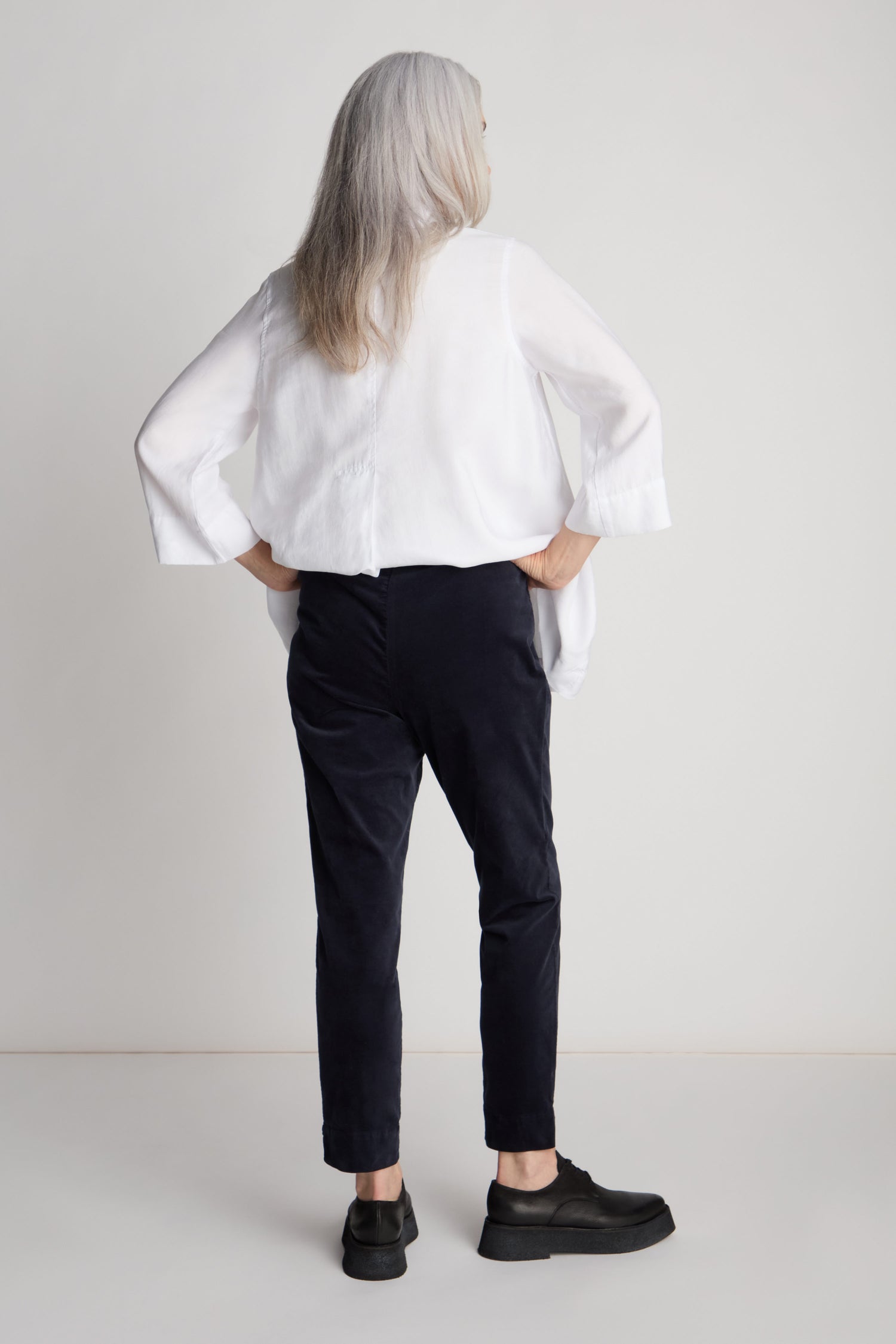 A person with long grey hair stands facing away, wearing a white top, Cotton Moleskin Slim Trouser—a true wardrobe staple—and black shoes, with hands in pockets.