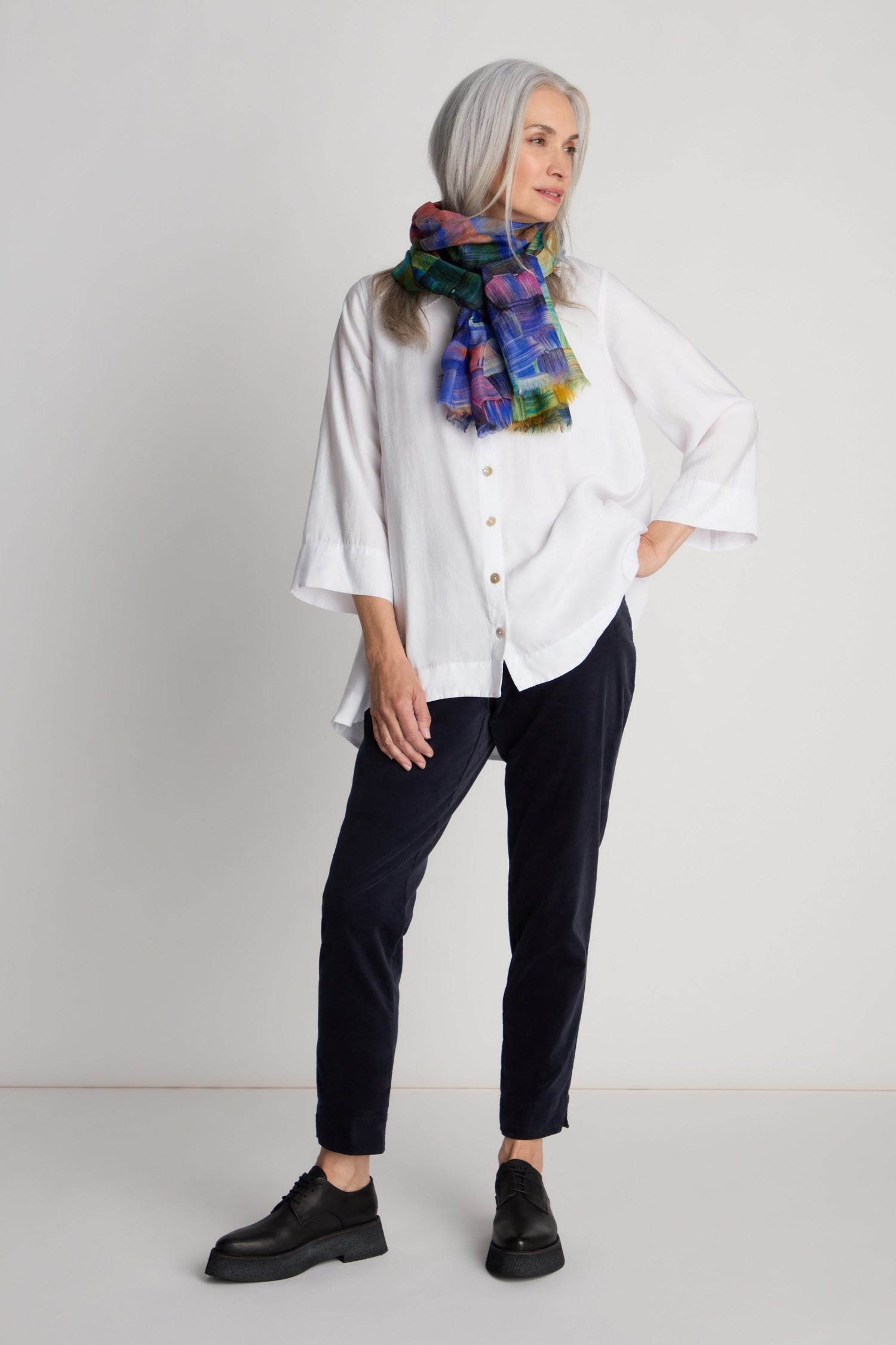 A woman with long gray hair is standing against a plain background, dressed in a white button-up blouse, Cotton Moleskin Slim Trousers, black shoes, and a colorful scarf.