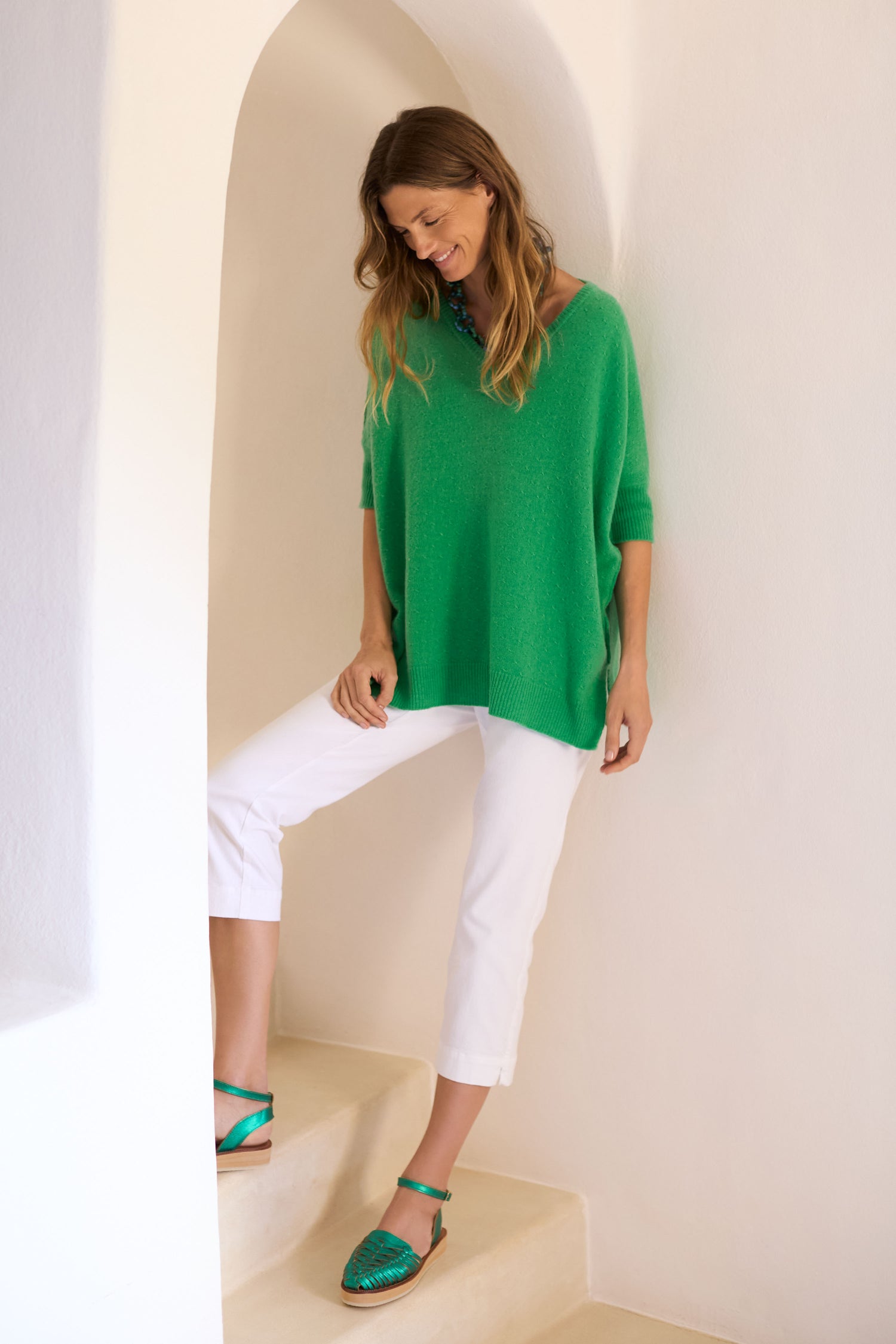 A woman wearing a green sweater and Stretch Cotton Slim Trouser stands on stairs against a white wall. She gazes down with a slight smile, perfectly showcasing these versatile wardrobe essentials.