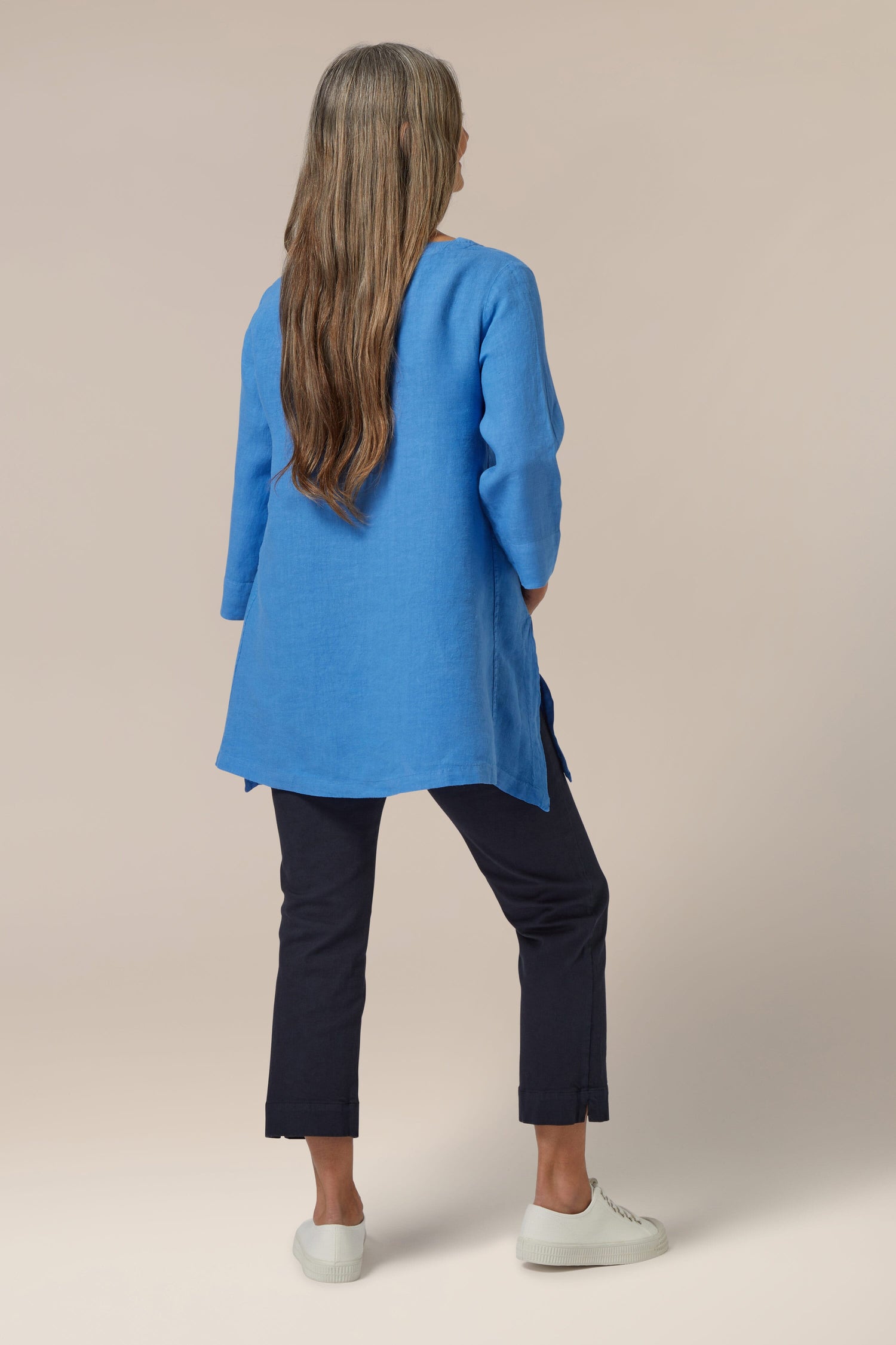 A woman standing with her back to the camera, wearing a blue tunic and navy Stretch Cotton Slim Crop Trousers with white slip-on shoes.