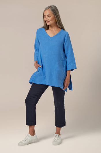 A woman in a versatile Stretch Cotton Slim Crop Trouser blue tunic and navy pants posing with her hands on her hips, showcasing an effortless layering staple for a slim silhouette.