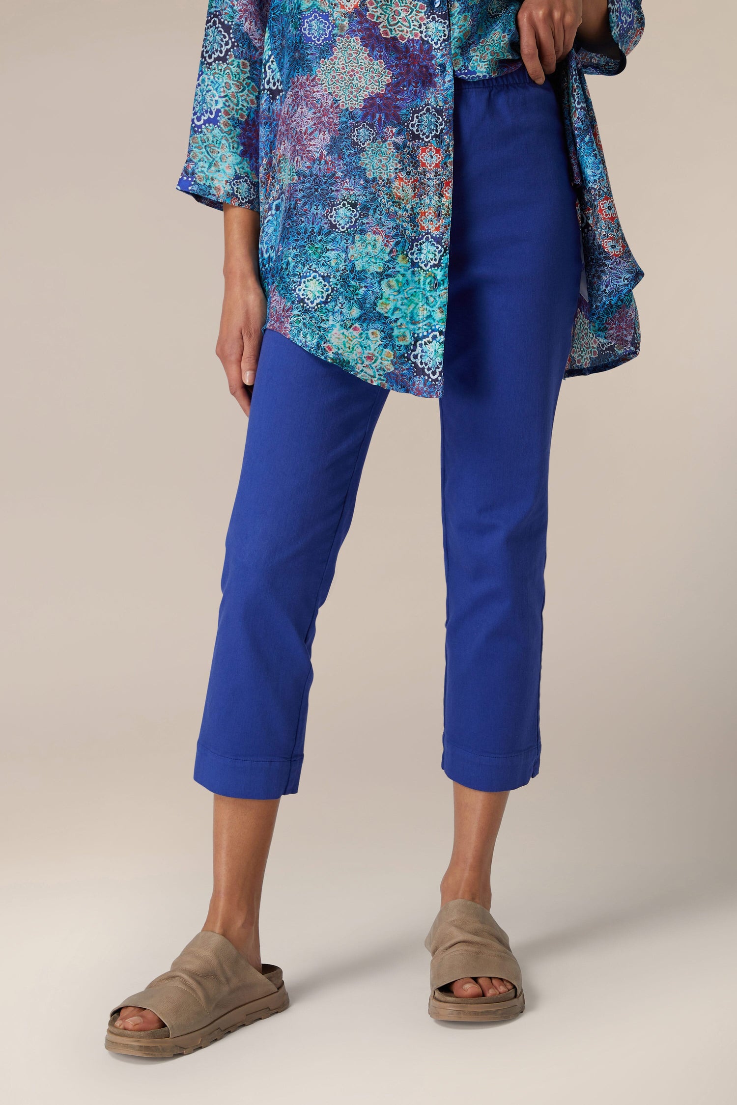 Woman wearing blue stretch cotton slim trousers and a floral blouse with taupe slide sandals standing against a neutral background.