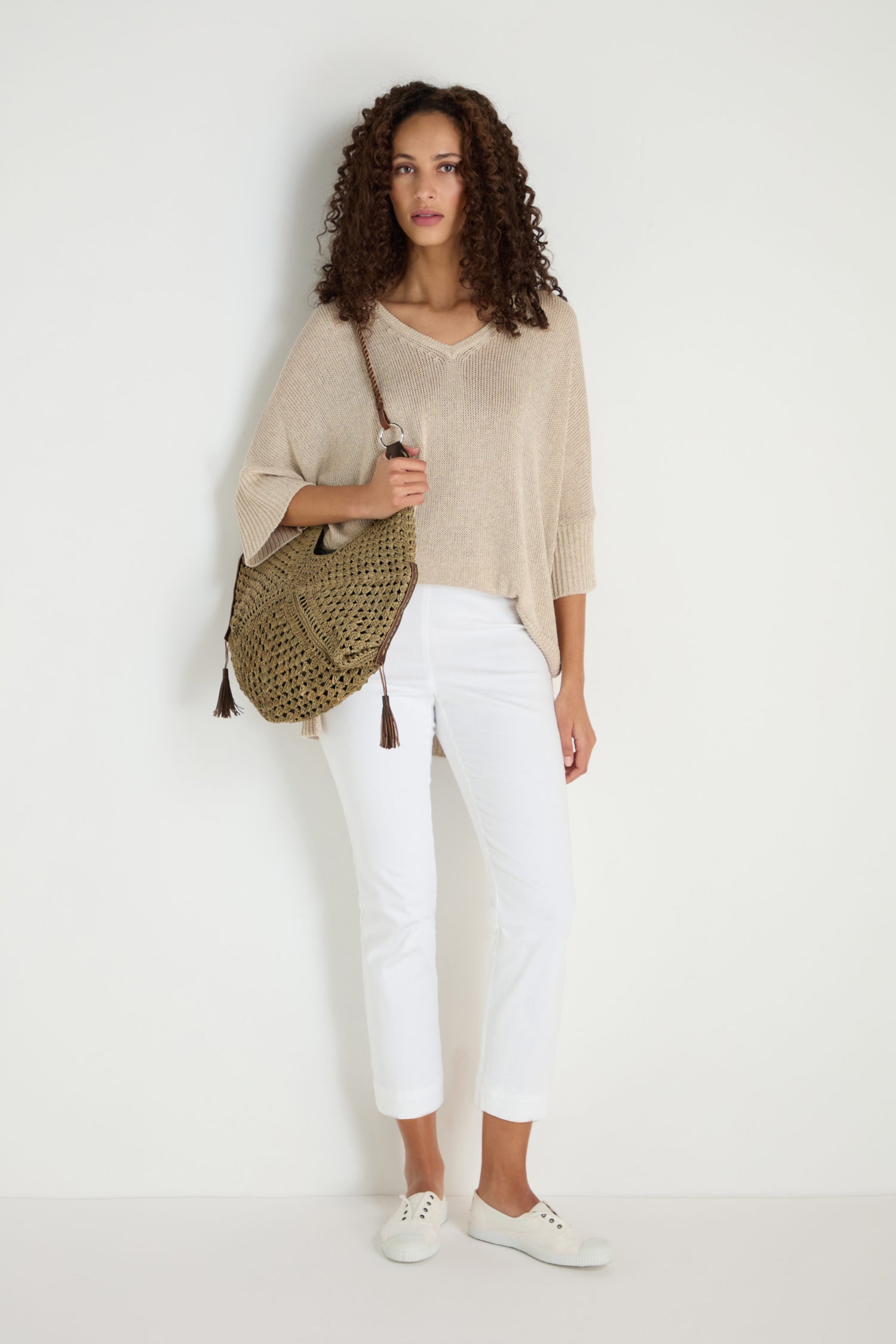 A woman stands against a white wall wearing ankle-grazing Stretch Cotton White Slim Trousers, a beige knit top, white sneakers, and carrying a woven shoulder bag—versatile staples for any wardrobe.