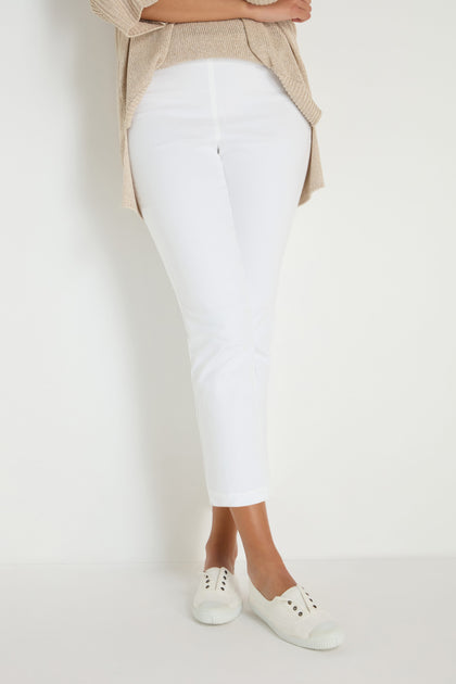 Wearing Stretch Cotton White Slim Trousers, a beige sweater, and white slip-on sneakers, the person stands against a plain white background—highlighting modern, versatile wardrobe essentials.