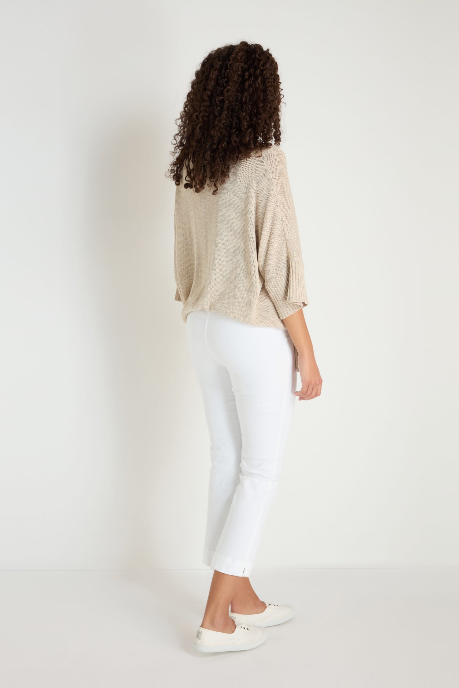Viewed from behind, a person with curly hair wears a beige sweater and Stretch Cotton White Slim Trousers with white shoes—versatile wardrobe staples—while standing against a plain white background.