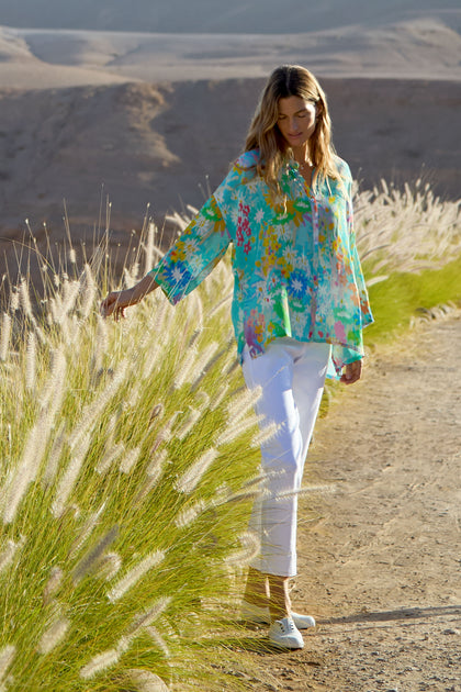 A woman in a colorful floral blouse and Stretch Cotton White Slim Trousers walks along a dirt path beside tall grass, with hills in the background.