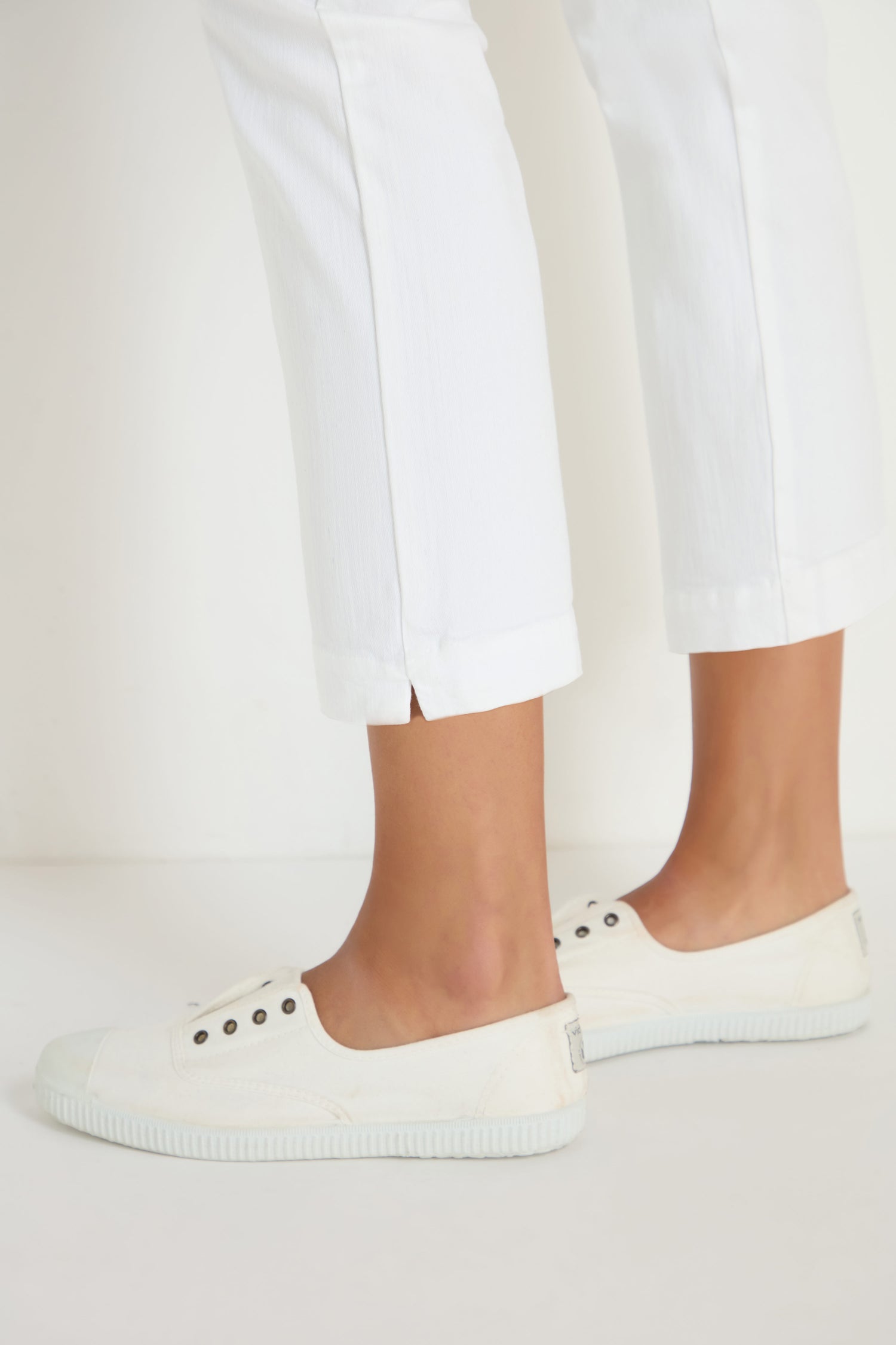 A person stands on a white surface with only their lower legs visible, wearing Stretch Cotton White Slim Trousers and slip-on sneakers—showcasing how these trousers are the ideal versatile wardrobe staple.