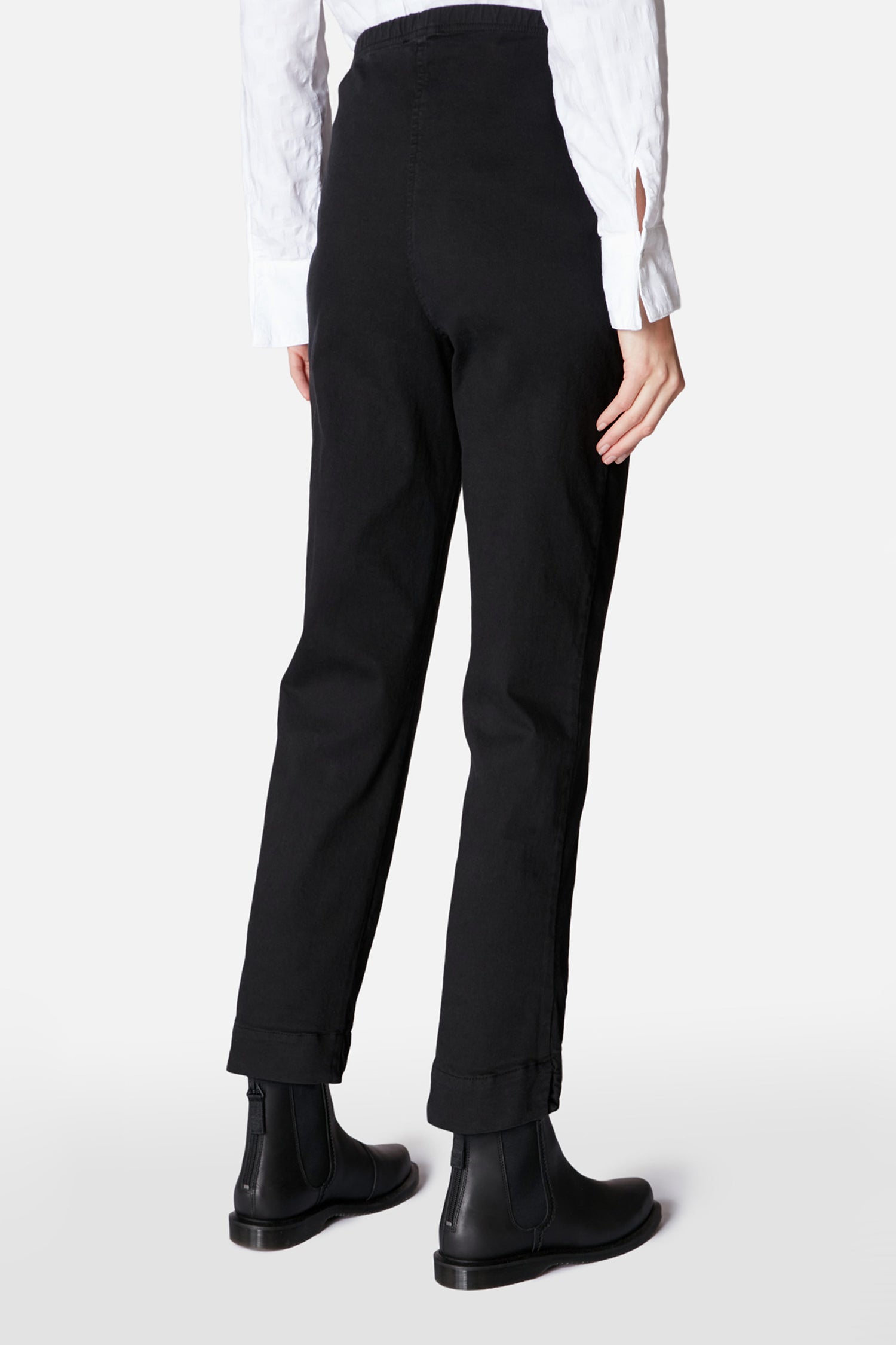 Person wearing Black Stretch Cotton Slim Trouser, black ankle boots, and a white long-sleeve shirt, shown from the back against a plain background.