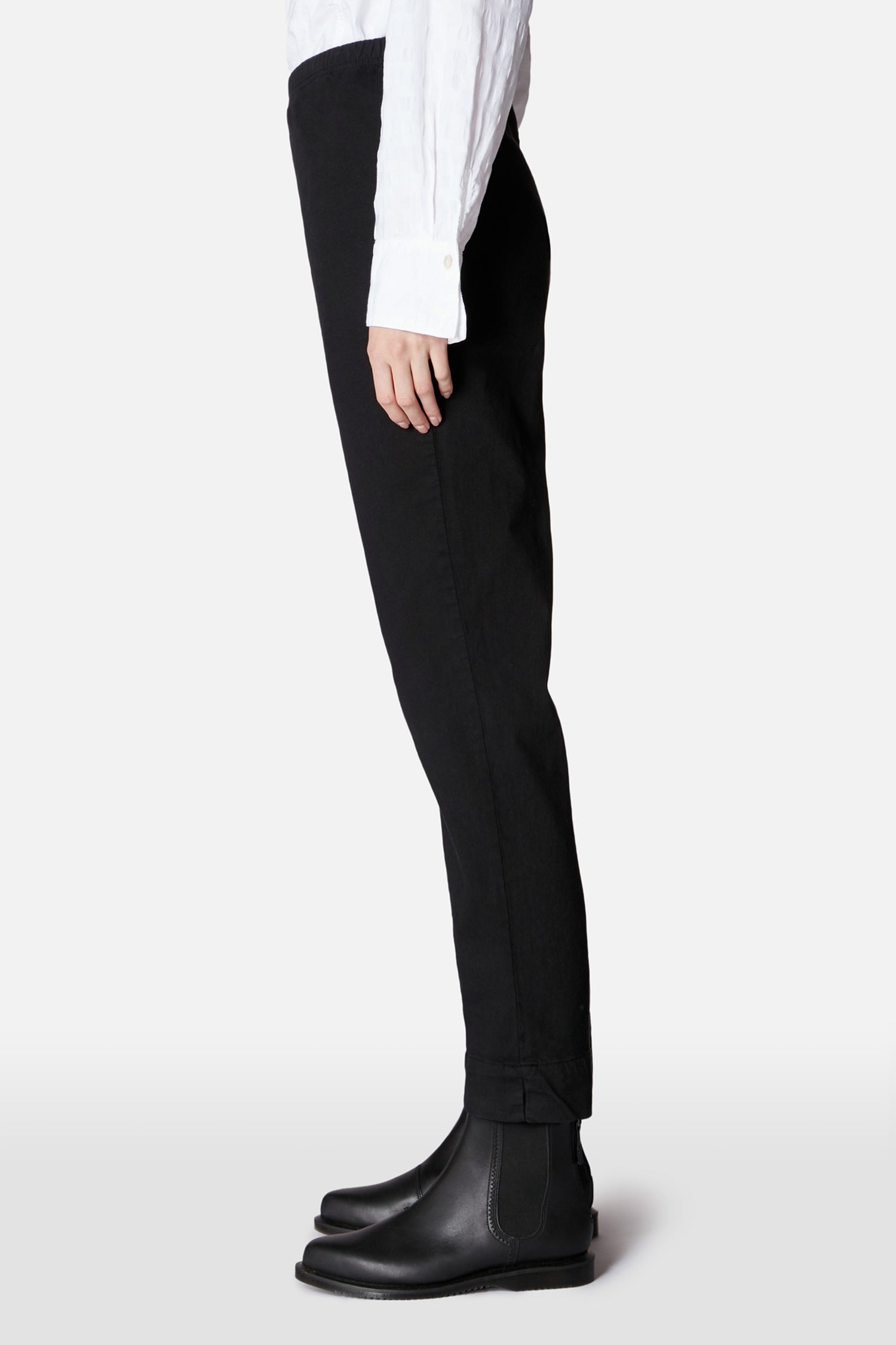 Person in profile wears a white long-sleeve shirt, Black Stretch Cotton Slim Trouser, and black Chelsea boots—versatile essentials against a plain light background.