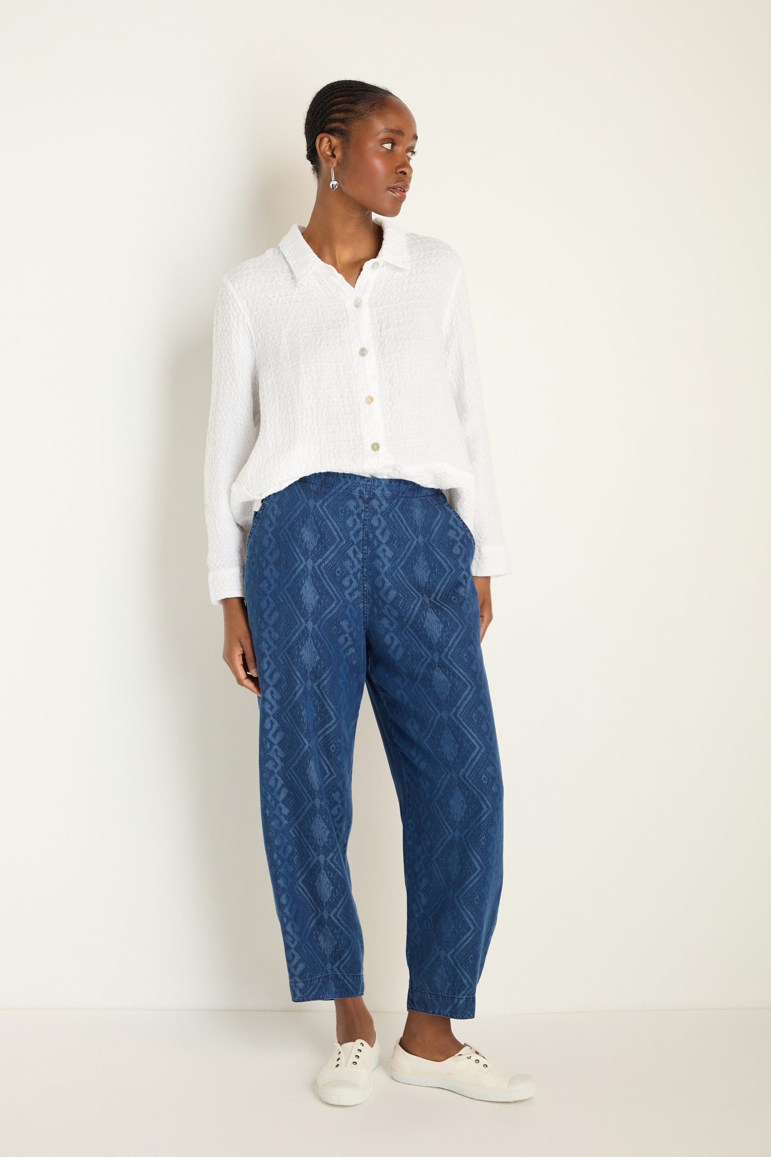 A person stands against a plain wall wearing a white textured button-up shirt, Light Denim Diamond Patchwork Print Bubble Trousers with an elasticated waistband, and white sneakers.