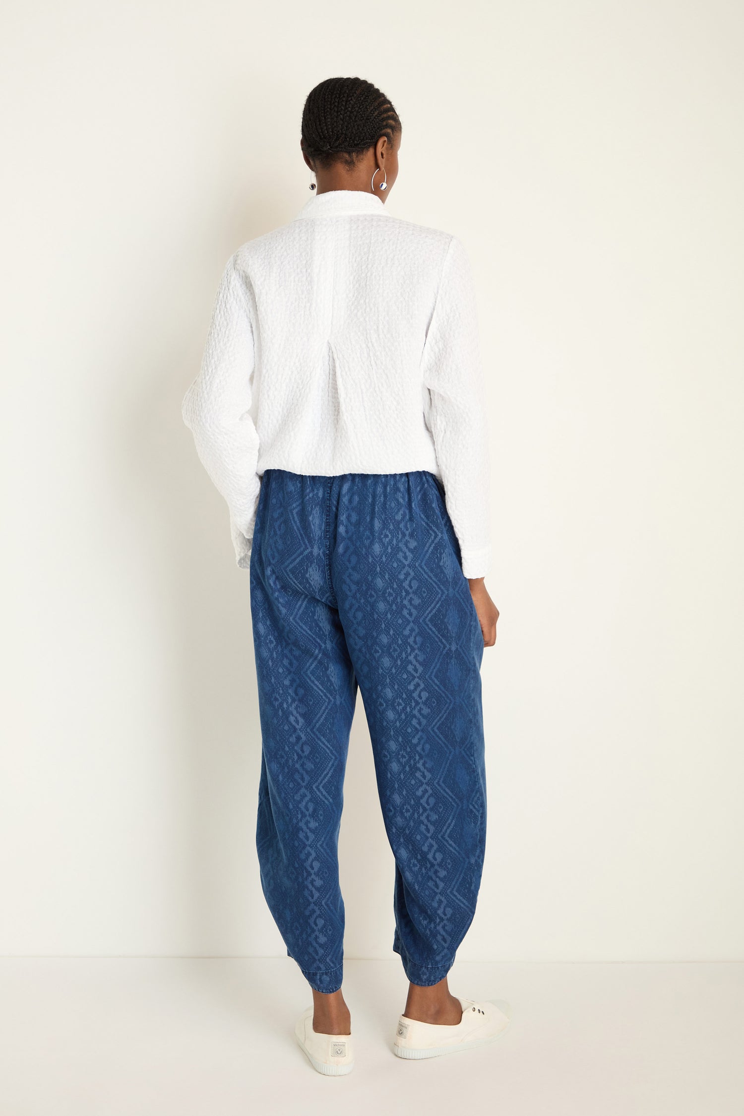 A person faces away from the camera, wearing a textured white shirt, Light Denim Diamond Patchwork Print Bubble Trousers with an elasticated waistband, and white shoes against a plain light background.