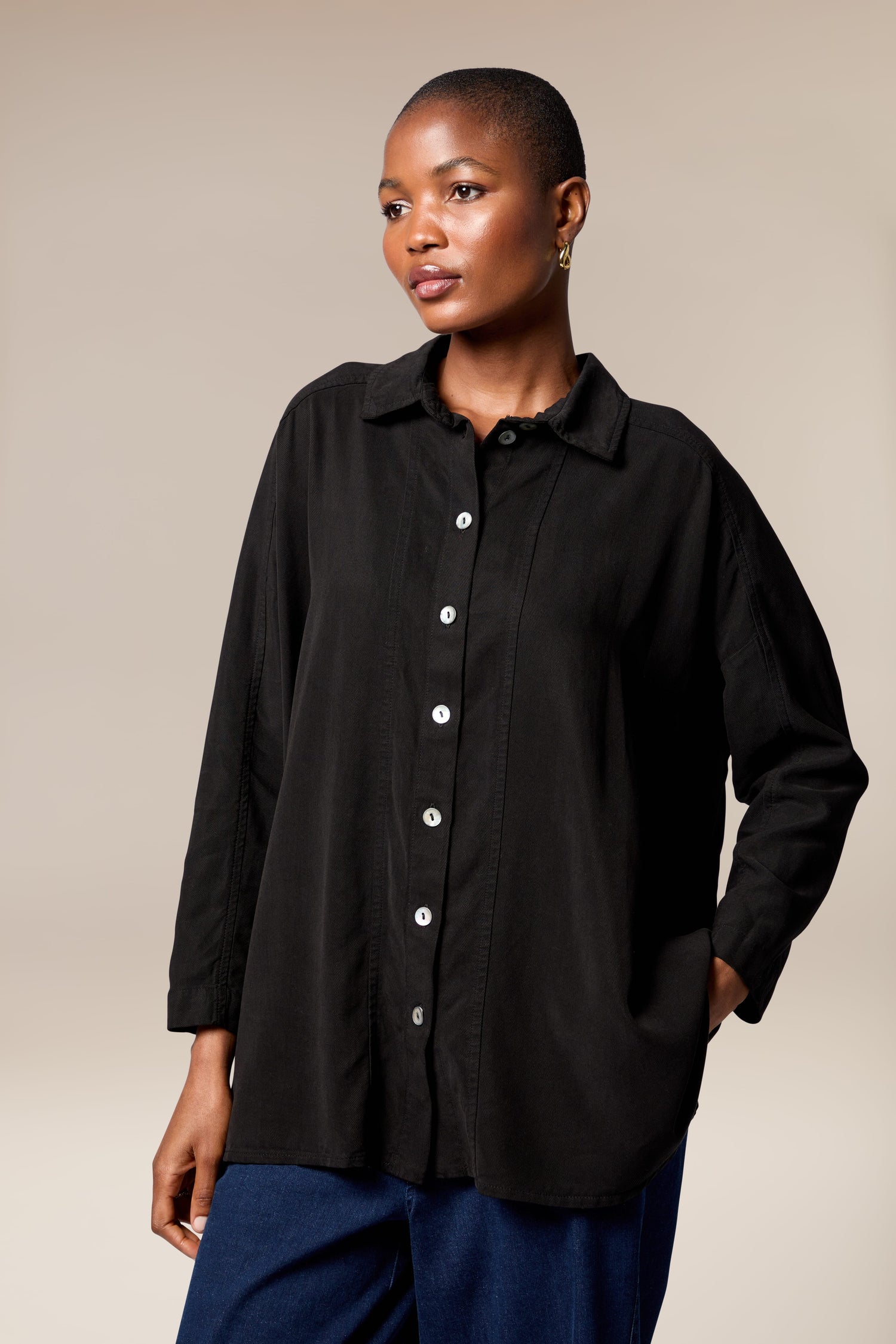 A person wears the Tencel Twill Boxy Shirt—a loose, boxy black button-up—paired with blue jeans, standing against a neutral background and looking slightly to the side.