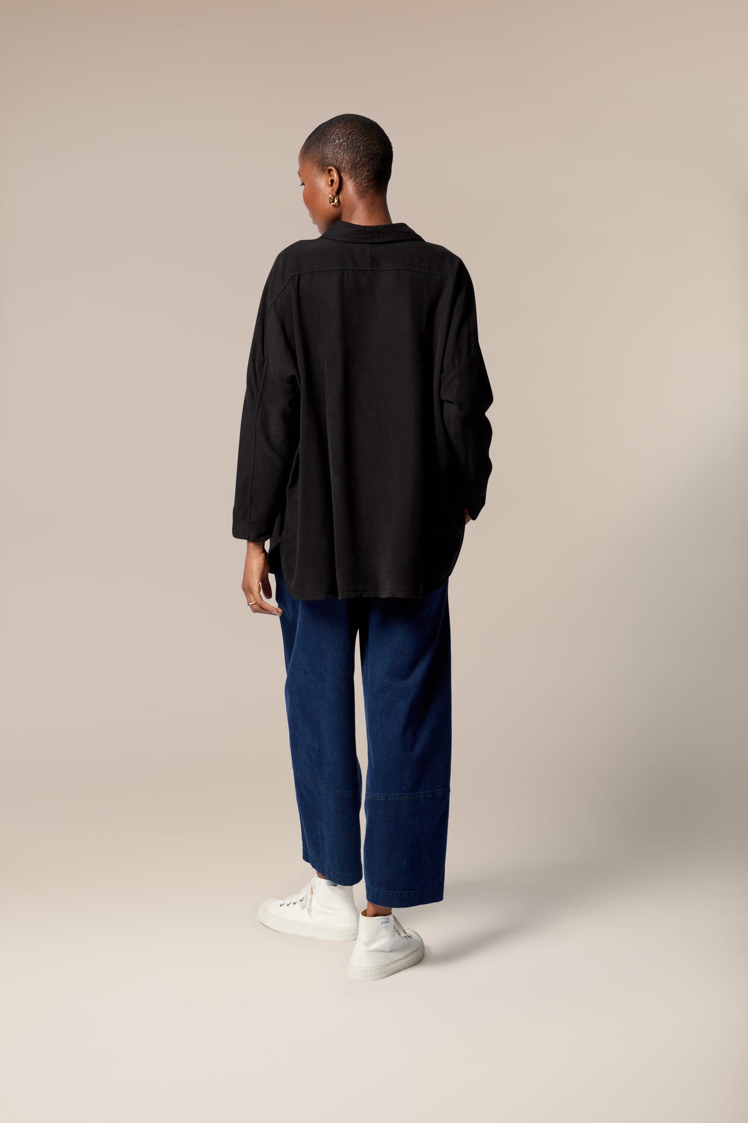 A person stands facing away, wearing the Tencel Twill Boxy Shirt in black, blue wide-leg pants, and white sneakers against a plain beige background.
