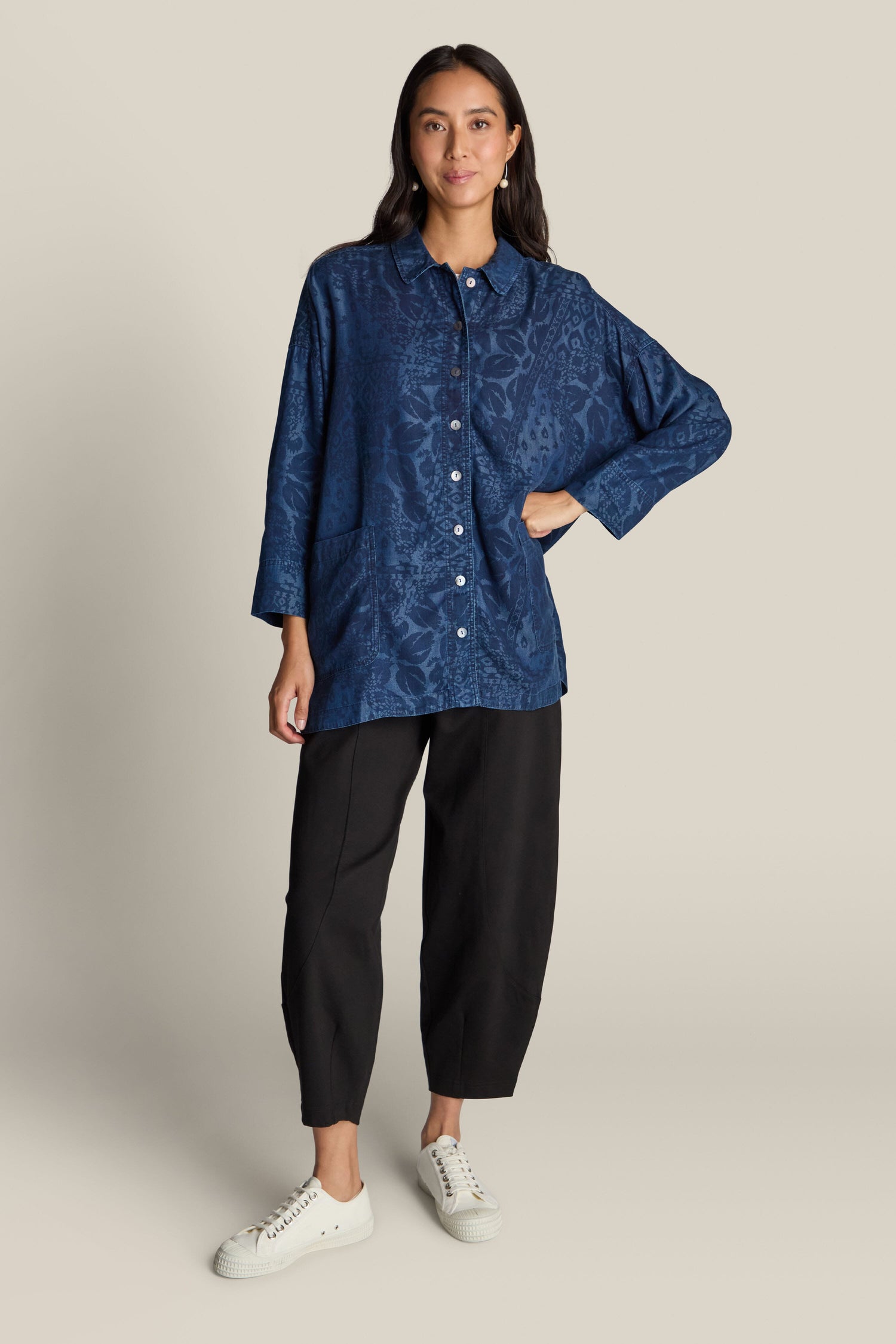 A person stands against a beige background wearing an Abstract Patchwork Print Shirt, featuring blue long sleeves and pockets, paired with black pants and white sneakers. The contemporary design of the shirt adds a stylish edge to the ensemble.