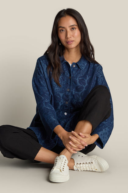 A person with long dark hair sits cross-legged wearing an Abstract Patchwork Print Shirt, black pants, and white sneakers against a plain background.