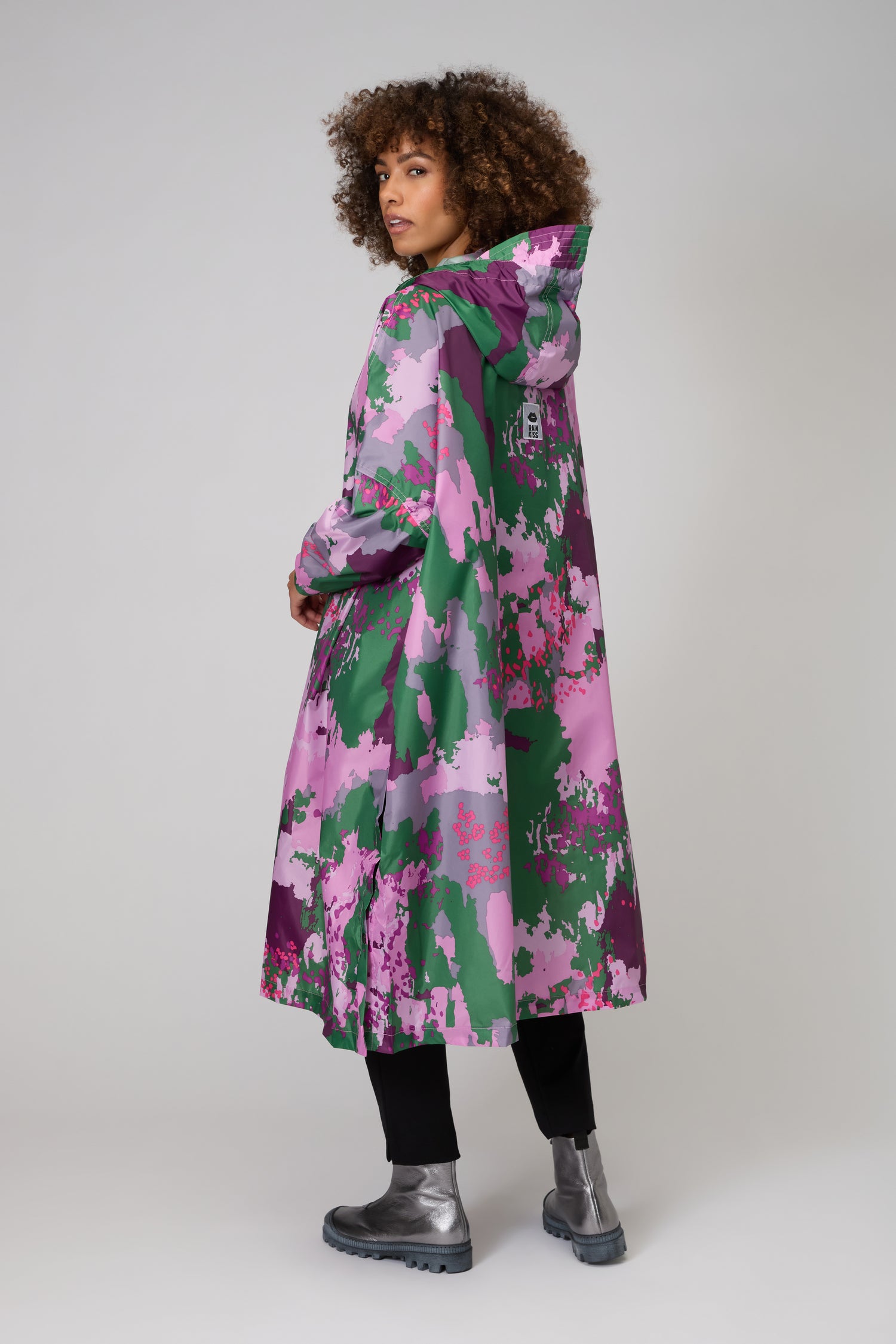 A person faces away, wearing the Digi Spring Poncho—a women’s hooded waterproof raincoat with a pink, green, and purple camo print—paired with black pants and silver boots.