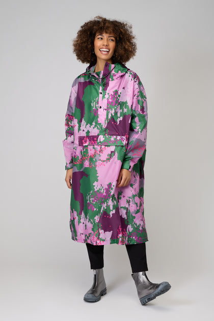 A person smiles in a bright, floral-patterned Digi Spring Poncho, paired with black pants and metallic boots, standing against a plain light background.