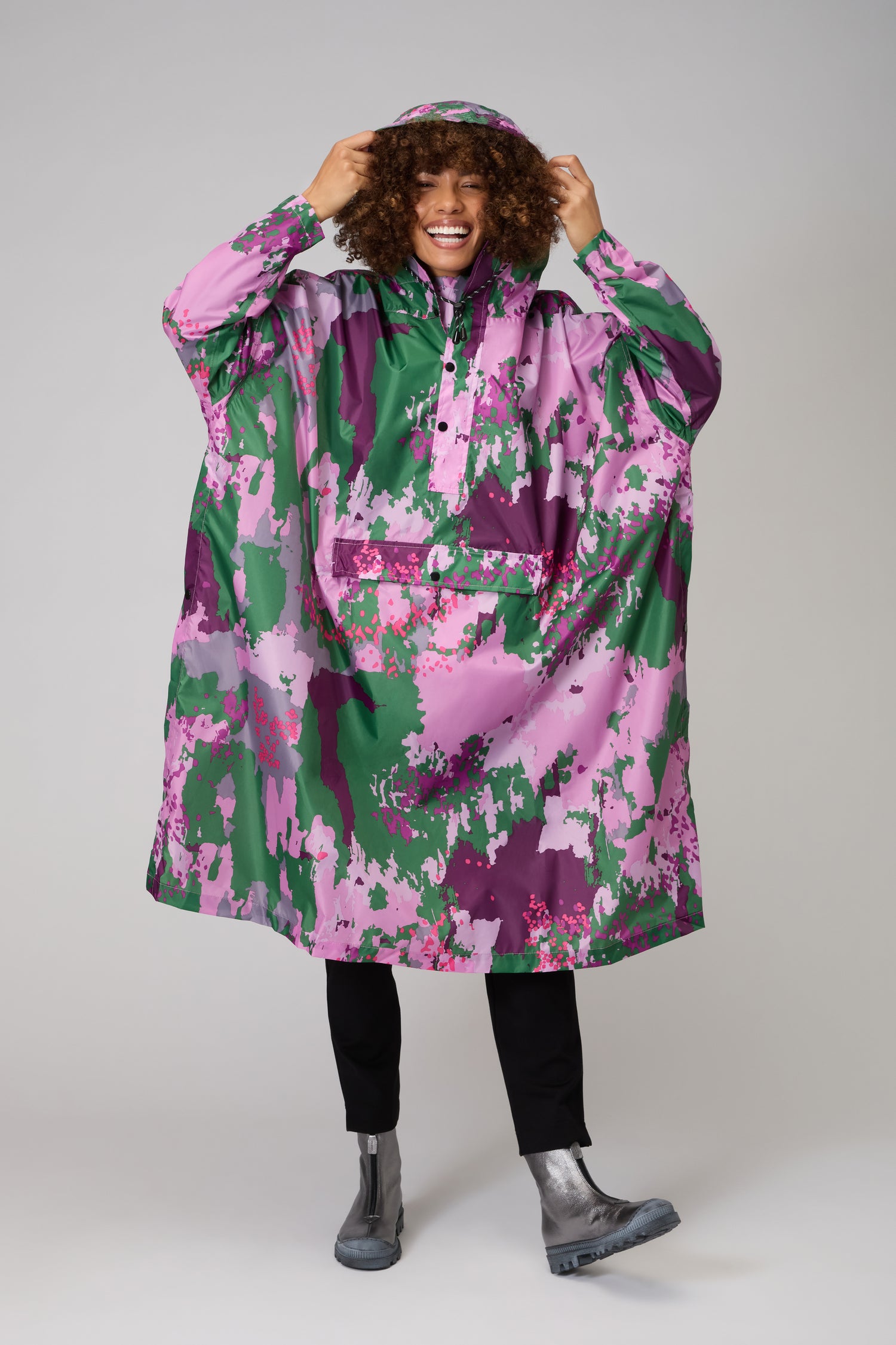 Person wears the Digi Spring Poncho, featuring a green, purple, and pink camo pattern in recycled polyester. Smiling and adjusting the hood, they show off this stylish, sustainable outerwear paired with black pants and gray boots.
