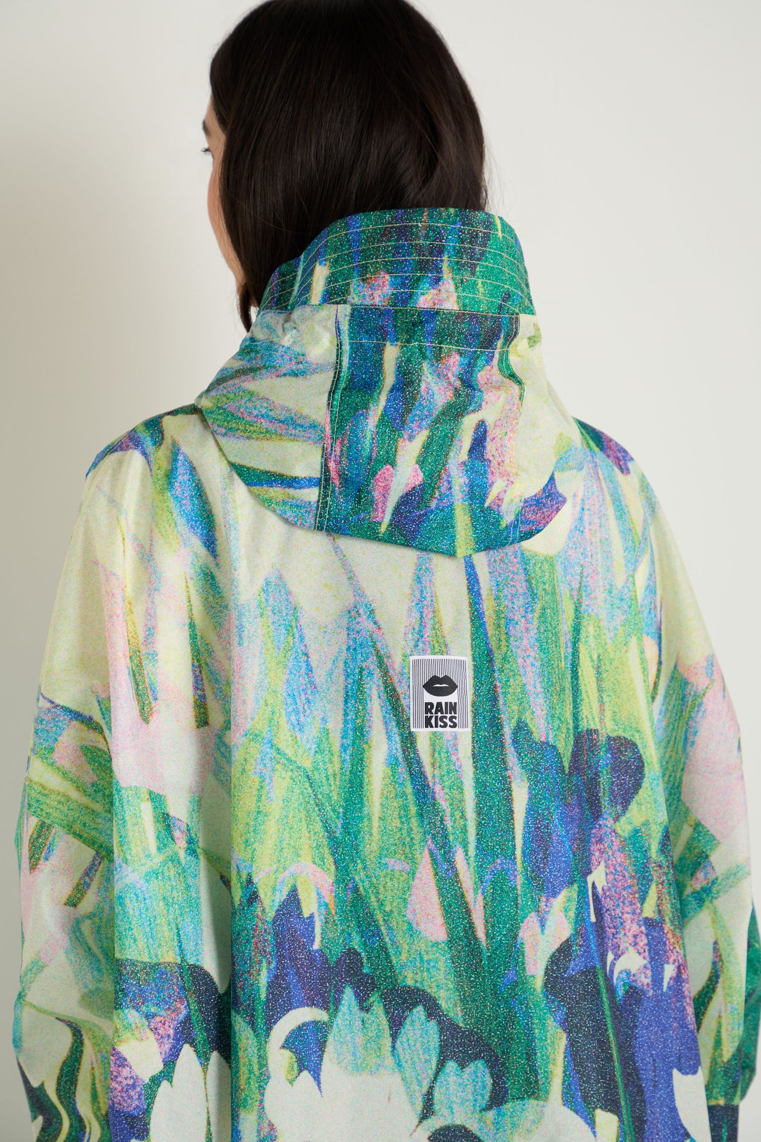 A person faces away from the camera, wearing the Lily Poncho—a colorful, patterned waterproof rain poncho with a hood; the "RAINS" brand label is visible on the back.