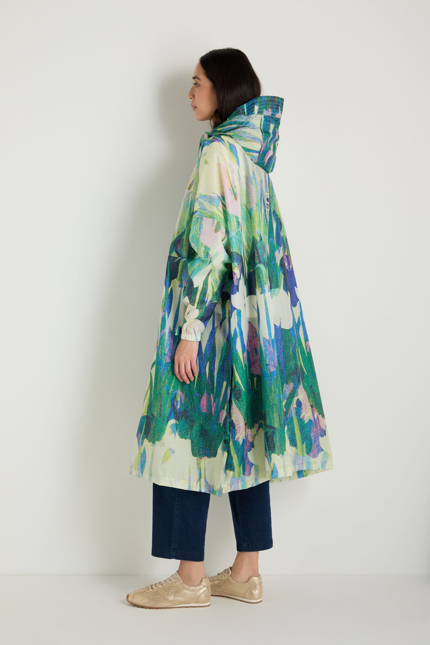 A person stands sideways against a plain white background wearing the Lily Poncho, a hooded, loose-fitting rain poncho with a colorful abstract floral pattern, paired with dark cropped pants and beige shoes.