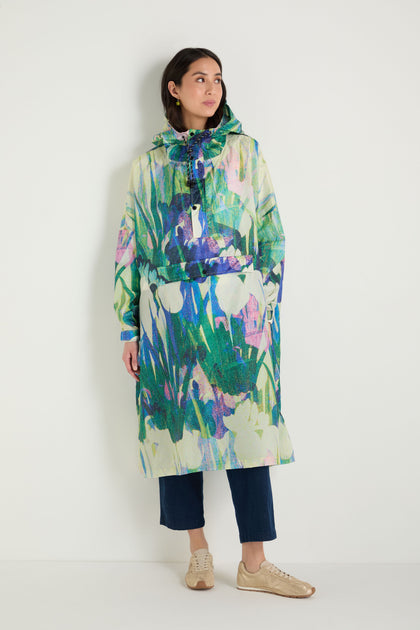 A woman stands against a white wall wearing the Lily Poncho, a long hooded rain poncho with an abstract floral pattern, paired with dark cropped pants and beige sneakers.
