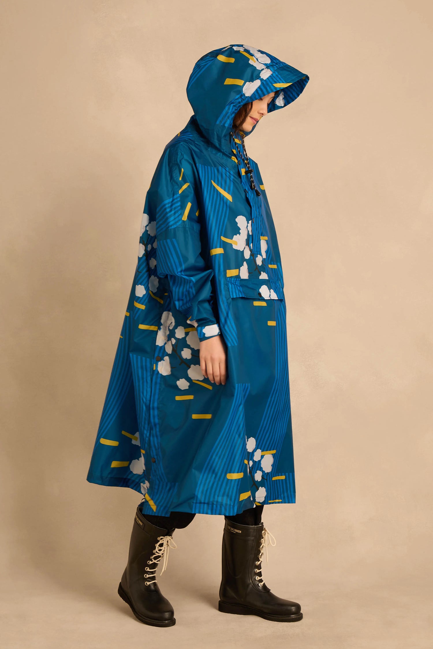 A person wears a Japanese Blue Blossom Poncho featuring blue fabric with white blossoms and yellow dash marks, paired with black lace-up rain boots.