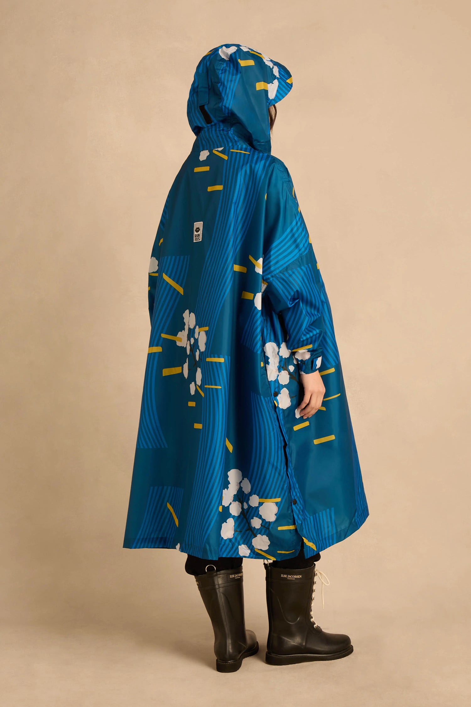 A person wears a Japanese Blue Blossom Poncho featuring white blossoms, yellow dashes, and curved lines, paired with black mid-calf rain boots.