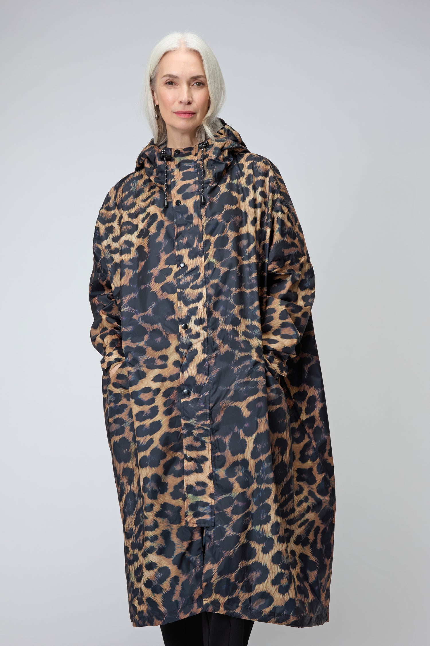 A person with long white hair wears an oversized, hooded Dark Leopard Rain Coat made from recycled polyester, standing against a plain light gray background.