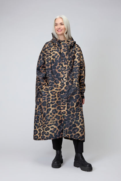 A person with long gray hair wears the Dark Leopard Rain Coat, made from recycled polyester, along with black pants and boots, standing against a plain light gray background.