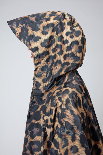A person in profile wears the Dark Leopard Rain Coat, a hooded poncho crafted from recycled polyester with a brown and black leopard print, shown against a plain light background.