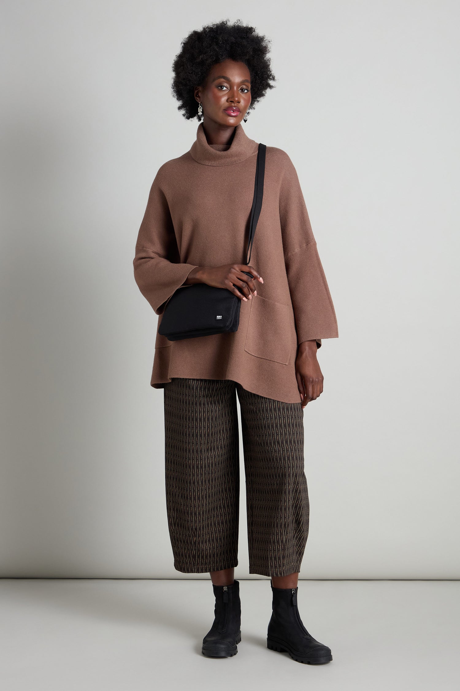 A person wearing a brown turtleneck sweater, patterned pants, and black boots stands against a plain background with the chic Recycled Carnaby Crossbody Bag slung over their shoulder.