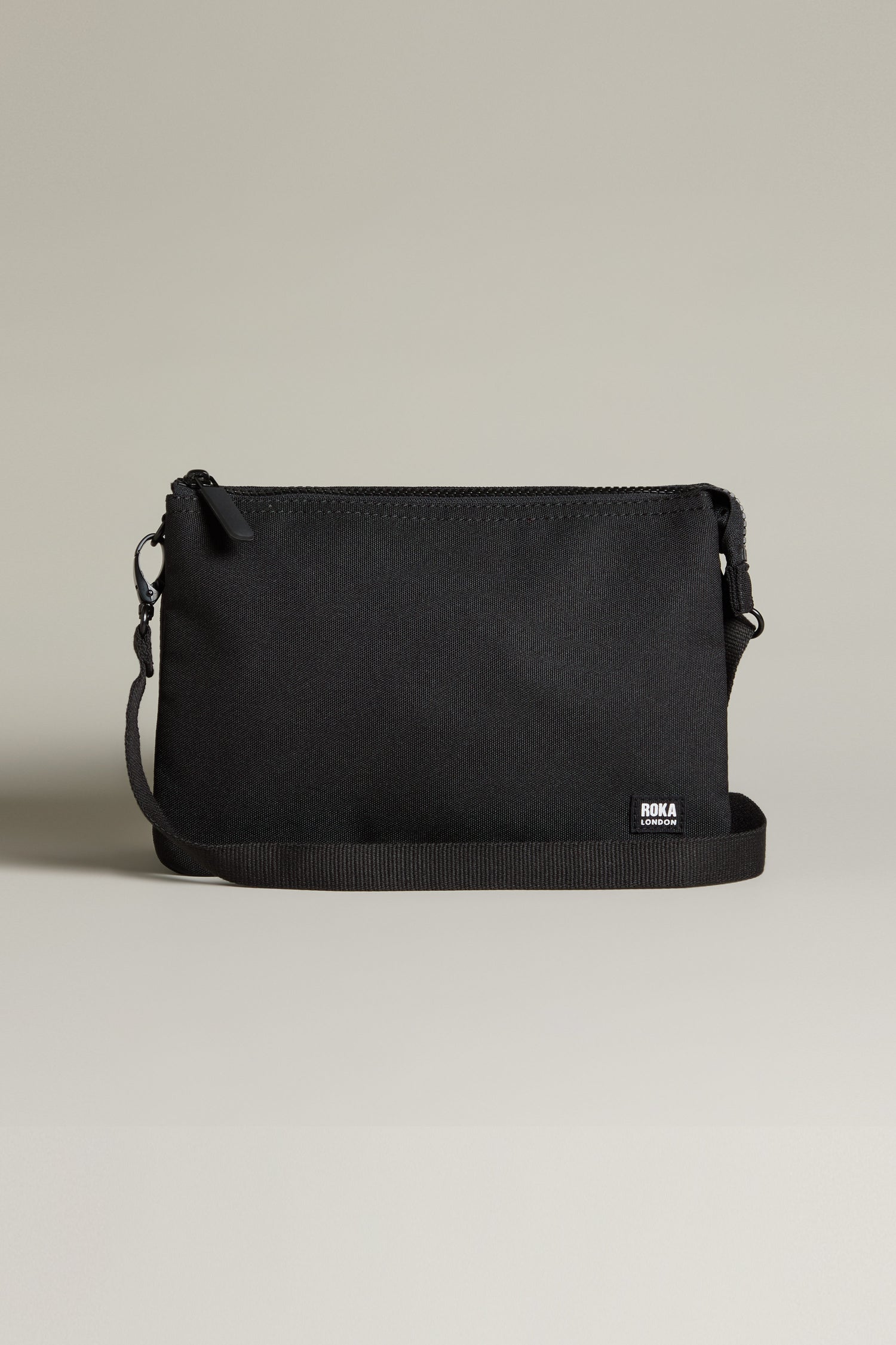 The Recycled Carnaby Crossbody Bag, made from recycled materials, features a sleek black design with a zippered top and adjustable strap, elegantly displayed against a neutral background.