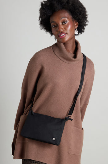 Person wearing a brown turtleneck sweater and carrying a sleek Recycled Carnaby Crossbody Bag, expertly crafted from sustainable materials, standing against a neutral background.