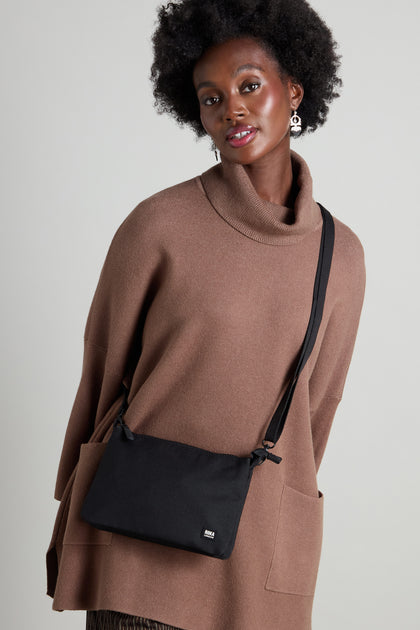 Person wearing a brown turtleneck sweater and carrying a sleek Recycled Carnaby Crossbody Bag, expertly crafted from sustainable materials, standing against a neutral background.